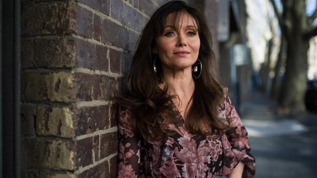 Essie Davis stands with one shoulder against a brick wall, staring off in the distance past the camera. Her left hand is on her hip. She is wearing a dark purple dress with floral motifs. She has big hoop earrings. Her long hair is down. 